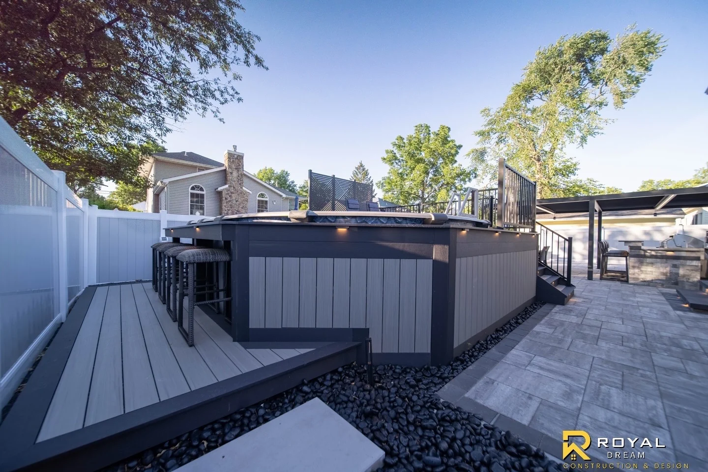 A raised deck and lower bar platform connect to the paver patio, with grey skirting and warm accent lights.