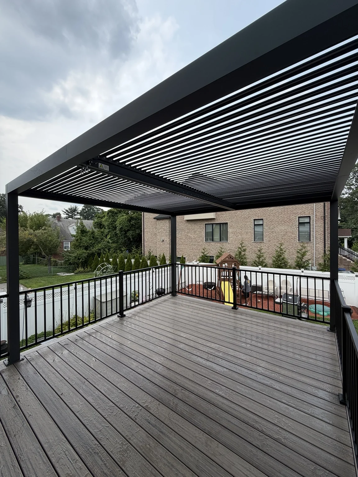 Grey deck boards beneath a black slatted pergola with open views through the perimeter railings.