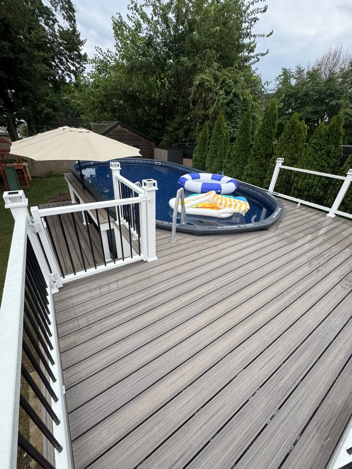 Grey deck boards curve around the pool with white posts, black balusters and an umbrella-shaded bar nearby.